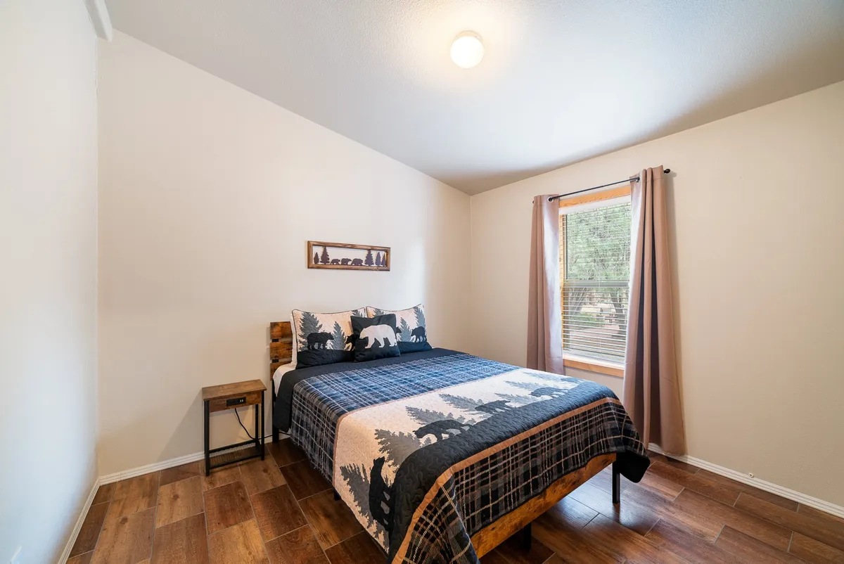 This welcoming bedroom features a bed with a bear-themed blanket, a wooden side table, and a window that lets in gentle, natural light. The decor is simple yet inviting, emphasizing relaxation and comfort.