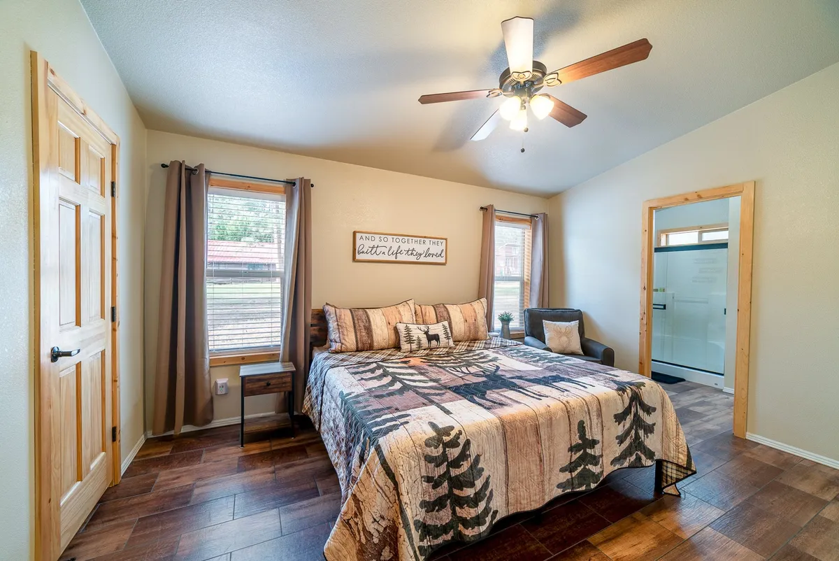 This image shows a spacious bedroom featuring a bed adorned with patterned bedding, wooden furniture, and abundant natural light streaming in from the windows. A decorative sign on the wall adds a homey touch. The room radiates calm and comfort, ideal for a restful night's sleep.