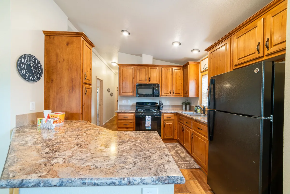 A welcoming kitchen with wooden cabinets, sleek black appliances, a granite countertop, and decorative elements such as a kitchen rug and wall clock. The space offers ample room for meal preparation and group interaction, reflecting a well-equipped and thoughtfully designed environment.