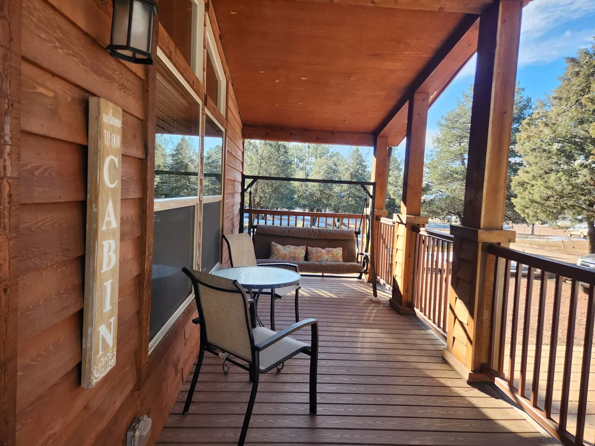 A charming porch swing and circular table rest under a wooden railing, with a friendly 'Welcome to Our Cabin' sign adding a personal touch. The swing faces outward, inviting couples to relax, watch sunsets, or savor a quiet conversation. Lush greenery borders the space, enhancing its intimacy.
