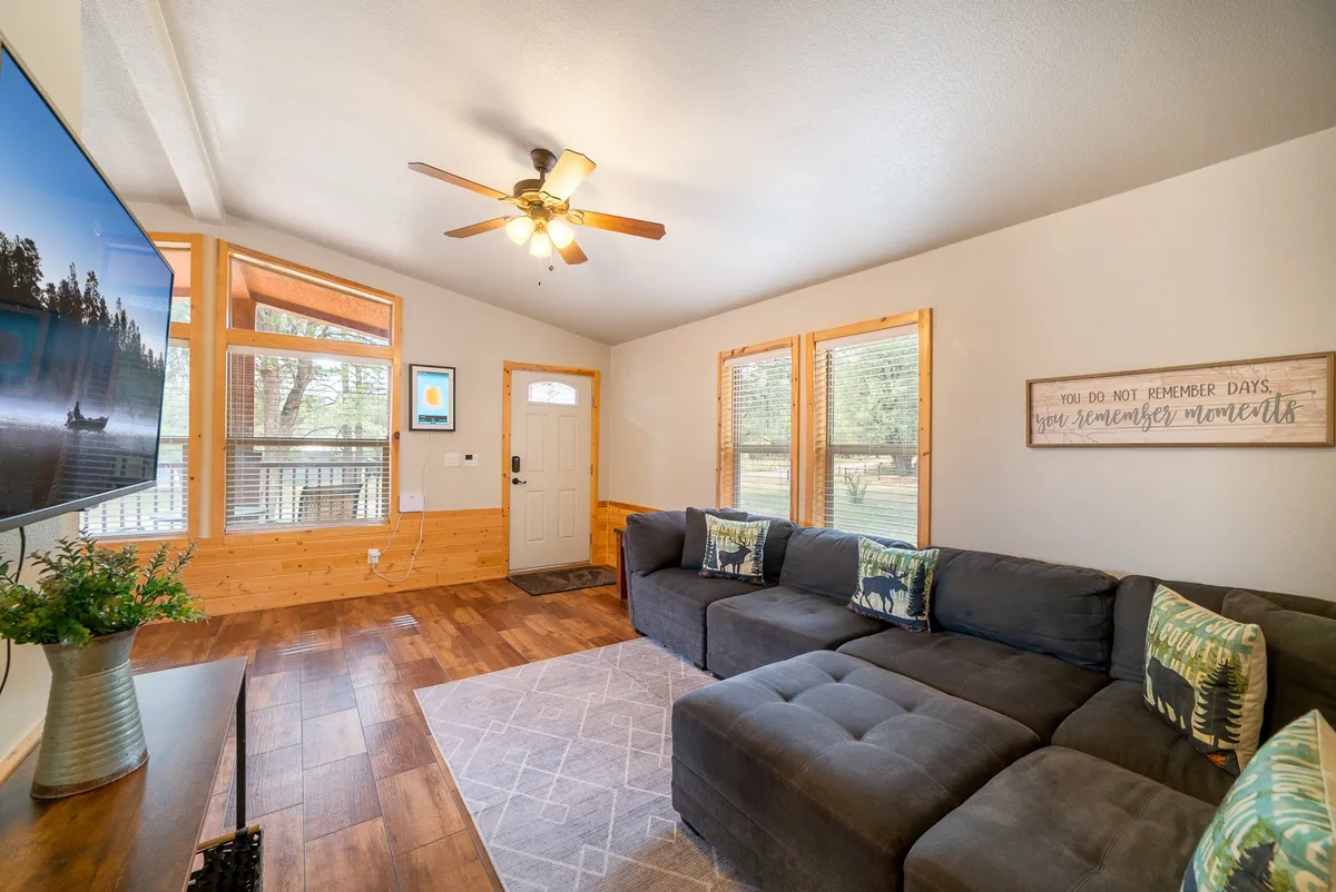 This image features a spacious living room with a sectional sofa, a TV, large windows letting in ample natural light, the entrance door, and decorative wall art. The scene radiates a cozy, inviting mood while clearly showcasing the living area and the large windows that frame the forest views outside.